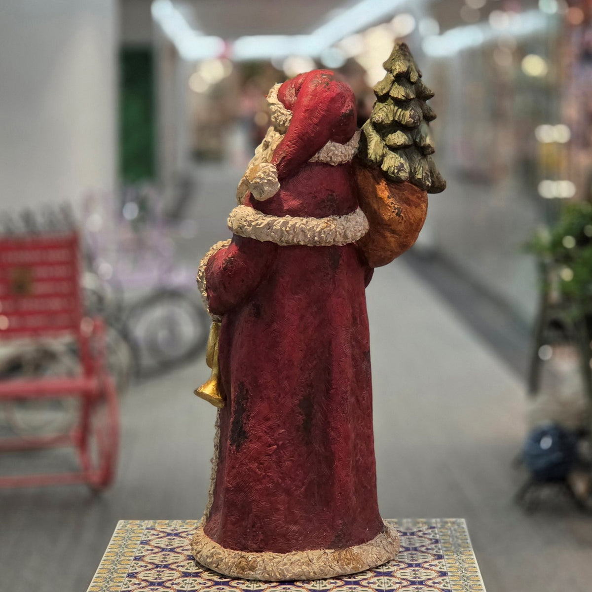 Rear view of a red-robed Santa figurine carrying a small Christmas tree and a gold horn.