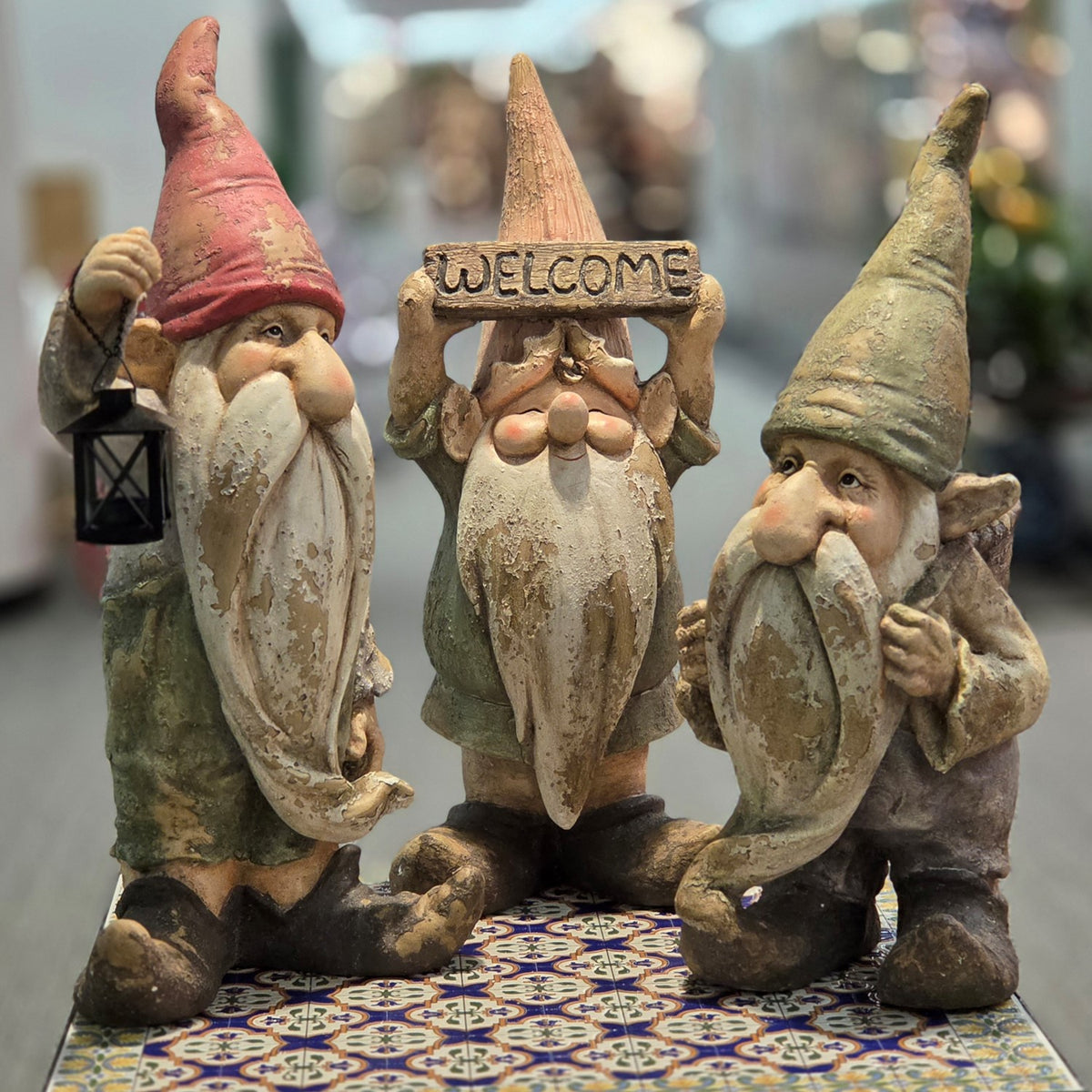 Three weathered garden gnomes stand together, with the center gnome holding a welcome sign overhead.