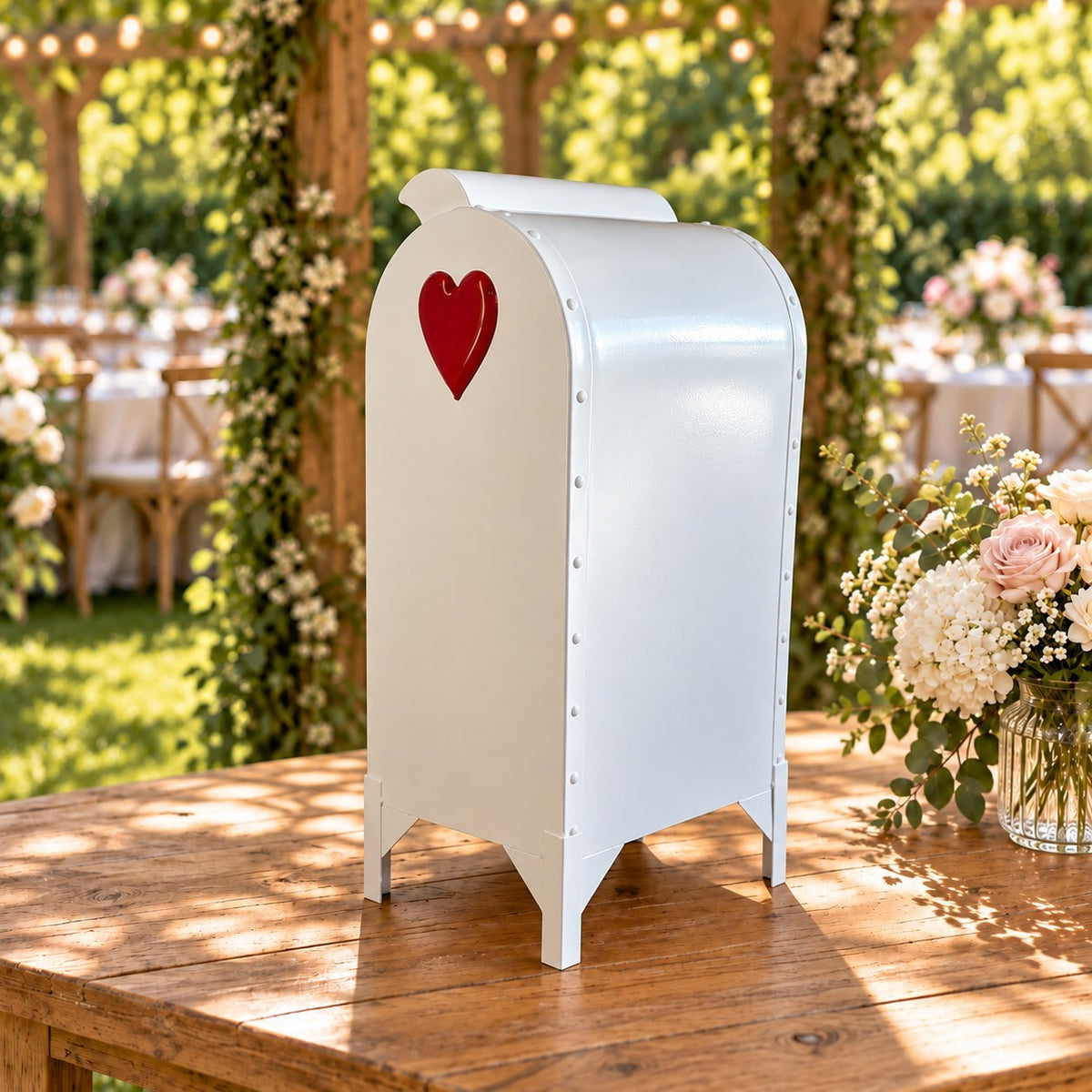 White curved-top mailbox with a red heart stands on a wooden table at an outdoor wedding setting.