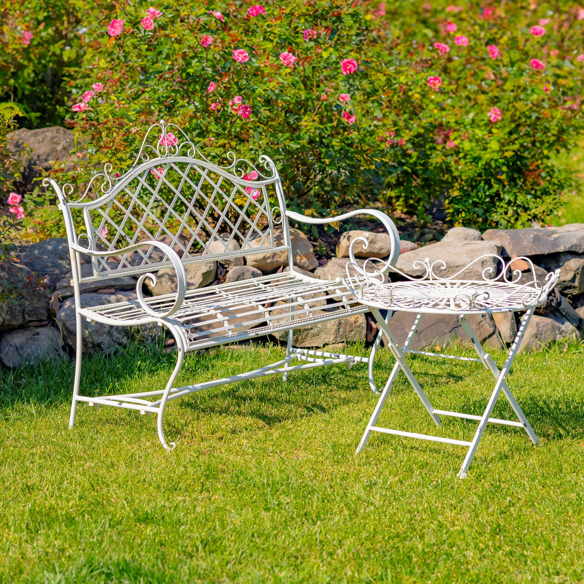 Ornate white metal table and matching lattice-back bench set on grass beside flowering rose bushes.