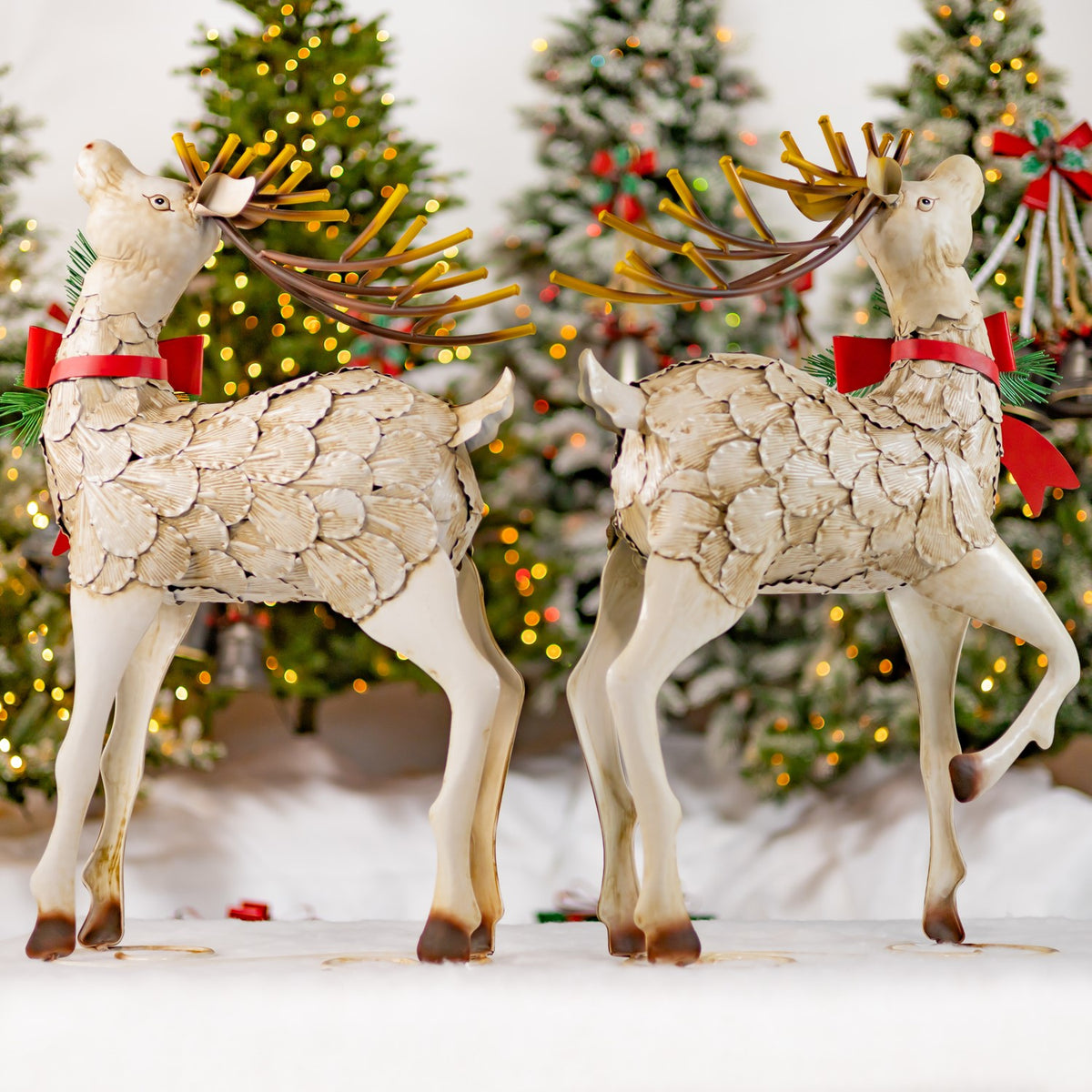 Two ivory reindeer figures with brown antlers and red pinecone bows stand in front of lit Christmas trees.