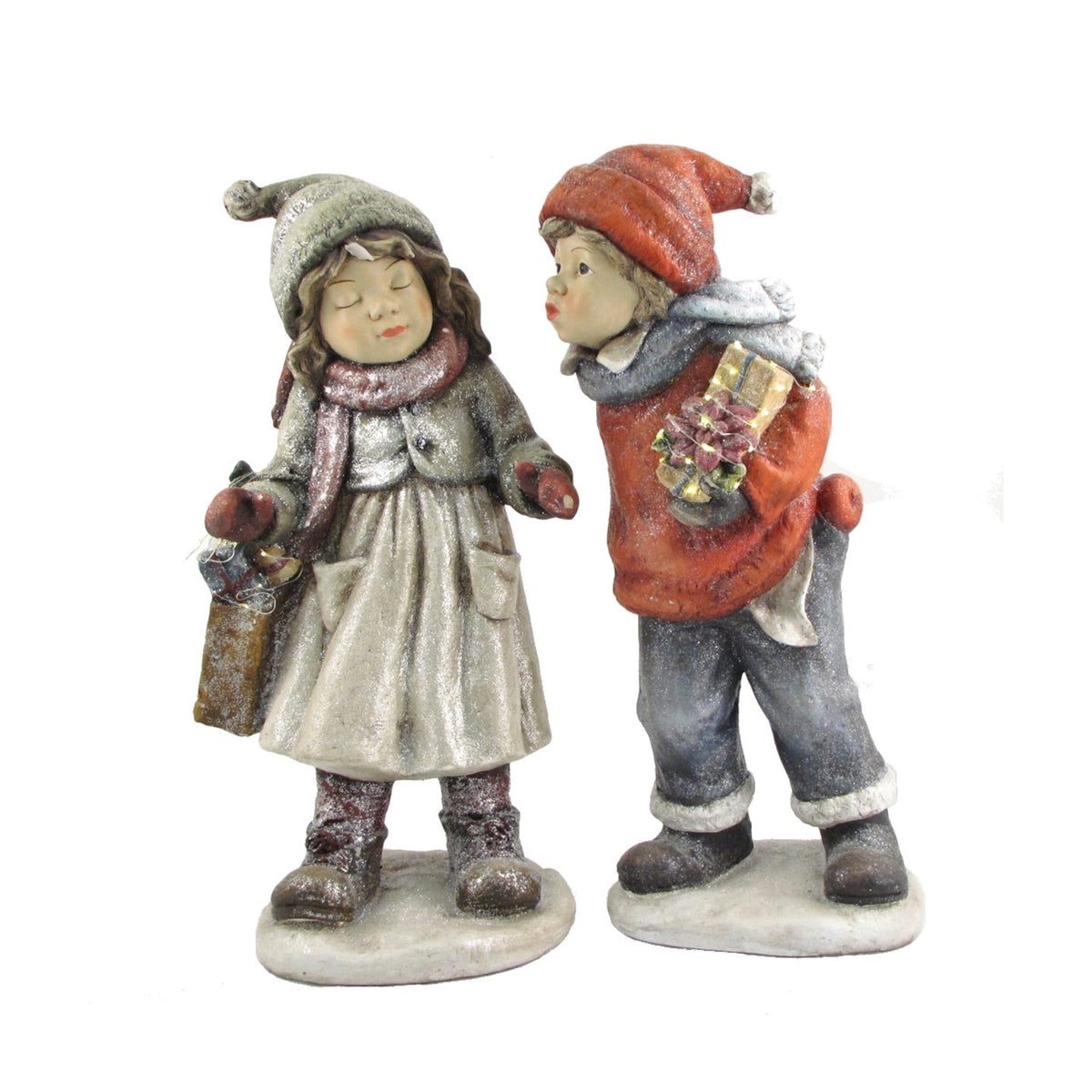 Two winter child figurines stand together, dressed in coats, hats and scarves and carrying gifts.