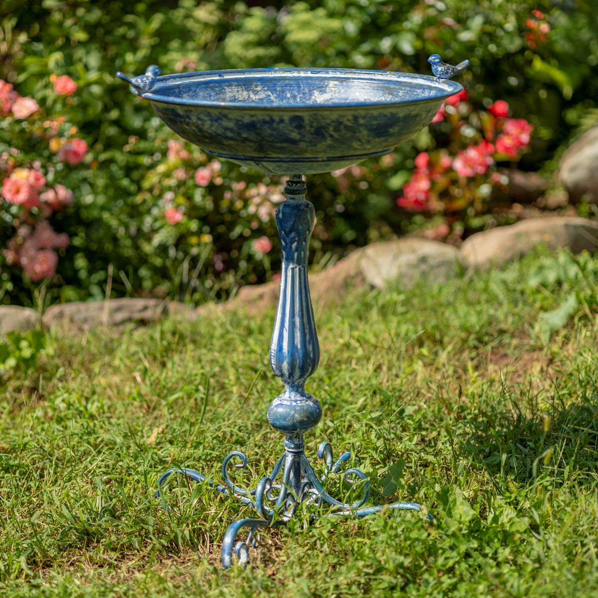 Blue ornate pedestal birdbath with small bird accents standing in a flower garden.
