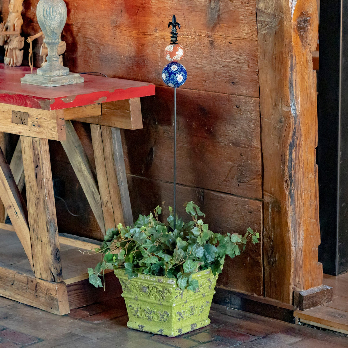 A decorative garden stake rises from a leafy planter beside a rustic wooden table.