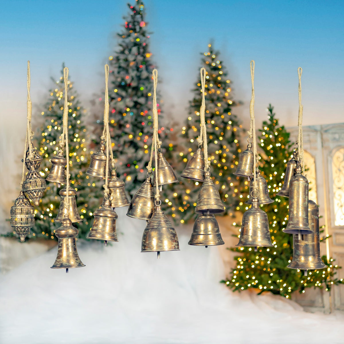 A group of assorted metal bells hangs in rows in front of decorated Christmas trees.