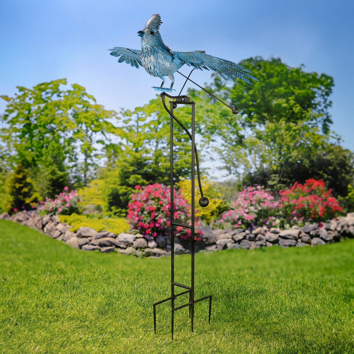 Full-height iron stake topped by a spread-wing blue jay, standing in a lawn among flowering shrubs.