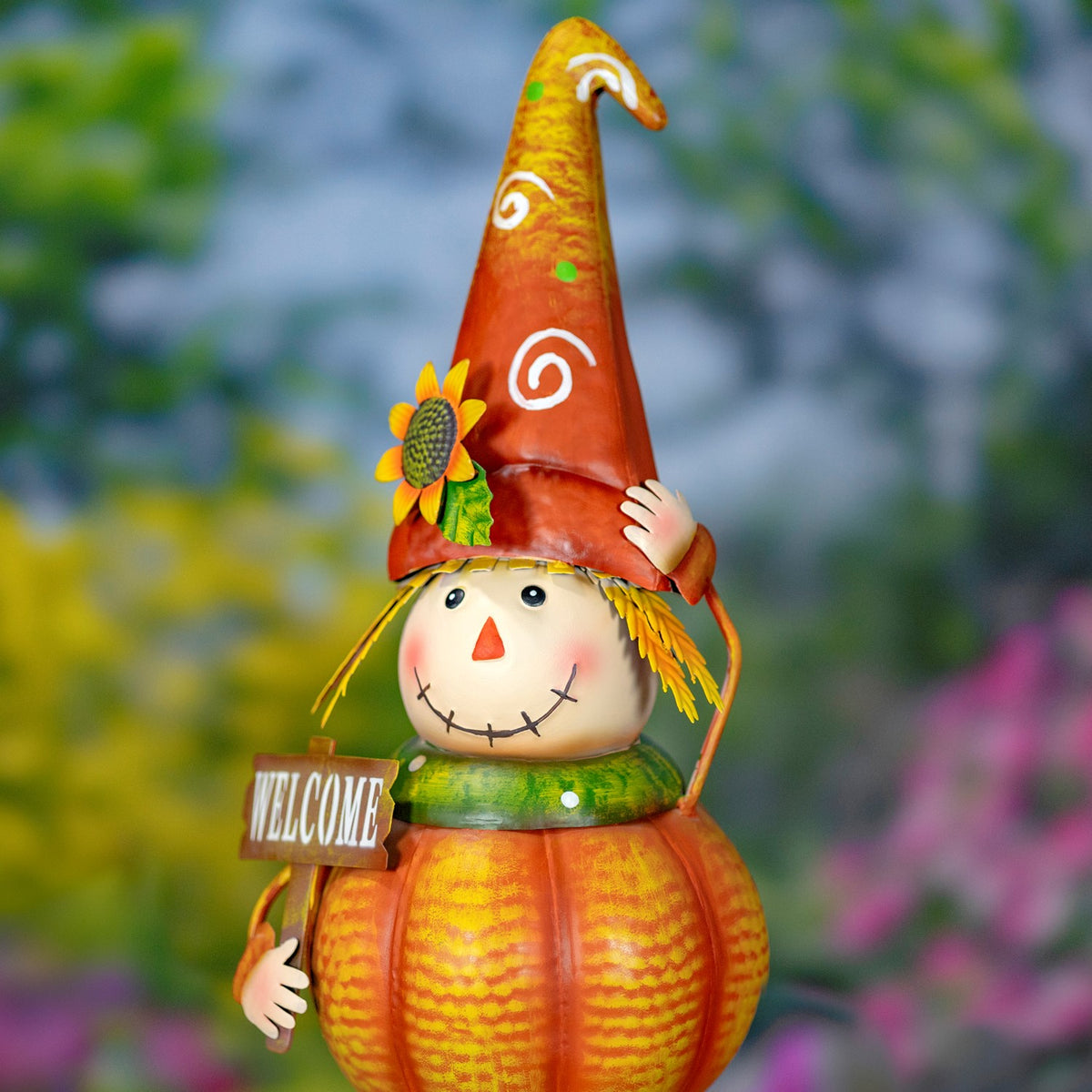 Close view of a smiling pumpkin scarecrow in a curled orange hat, raising one hand beside a sunflower.
