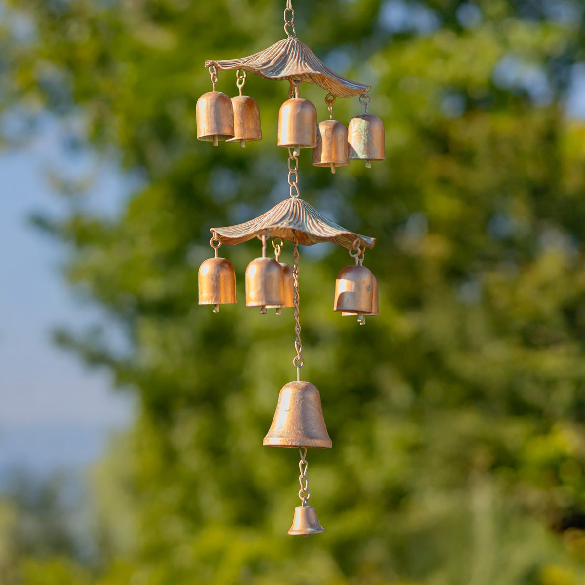Copper-colored hanging wind chime with tiered curved canopies and bells against leafy trees.