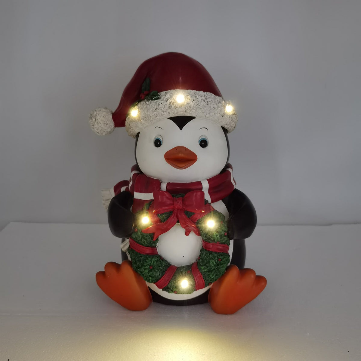 Seated penguin with a wreath, shown in a dim setting with lights glowing on its hat and body.