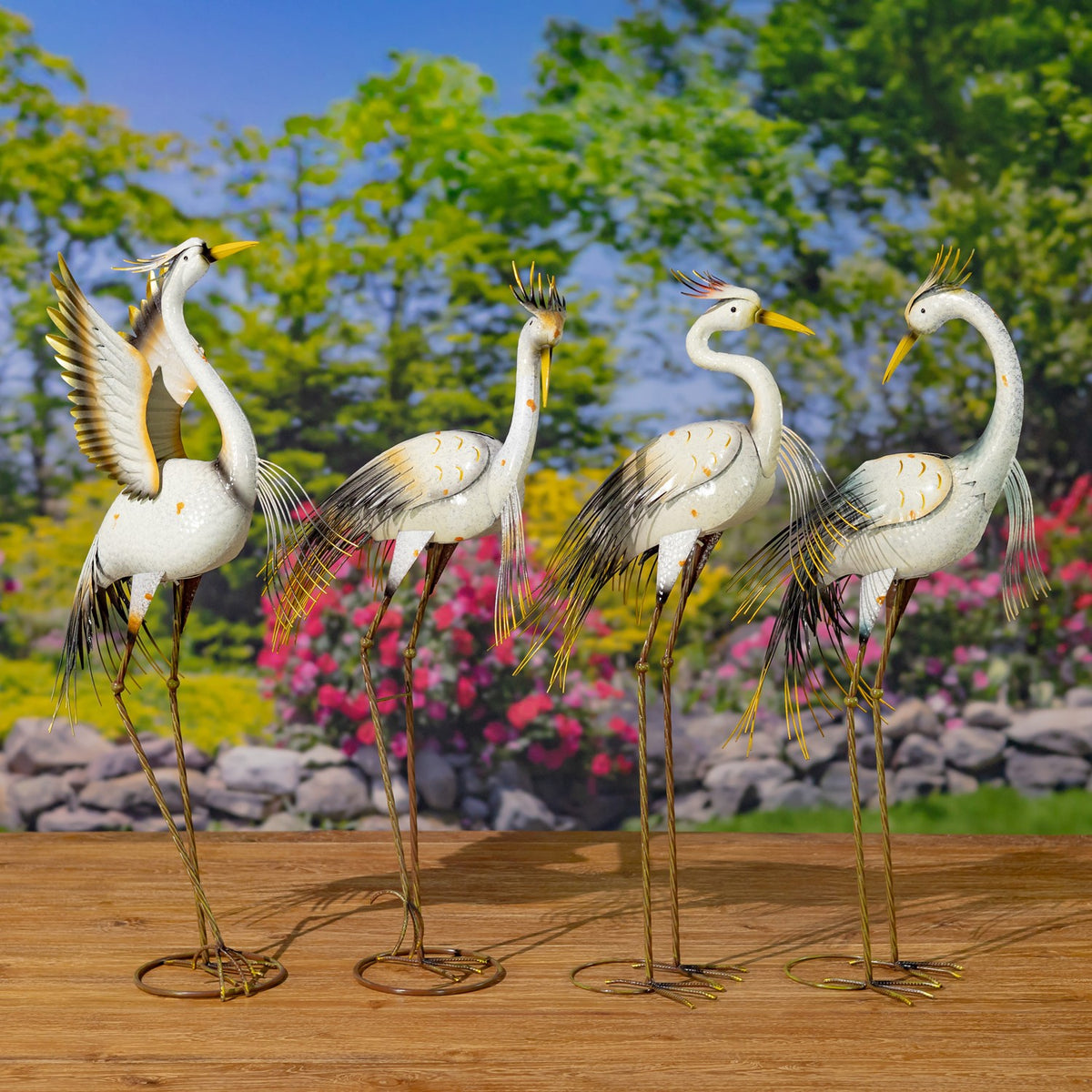 Four assorted metal heron figurines stand in a row on circular bases against a garden backdrop.