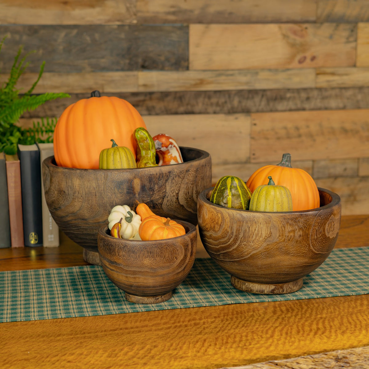 Three wooden bowls filled with decorative pumpkins and gourds on a tabletop against a plank wall.