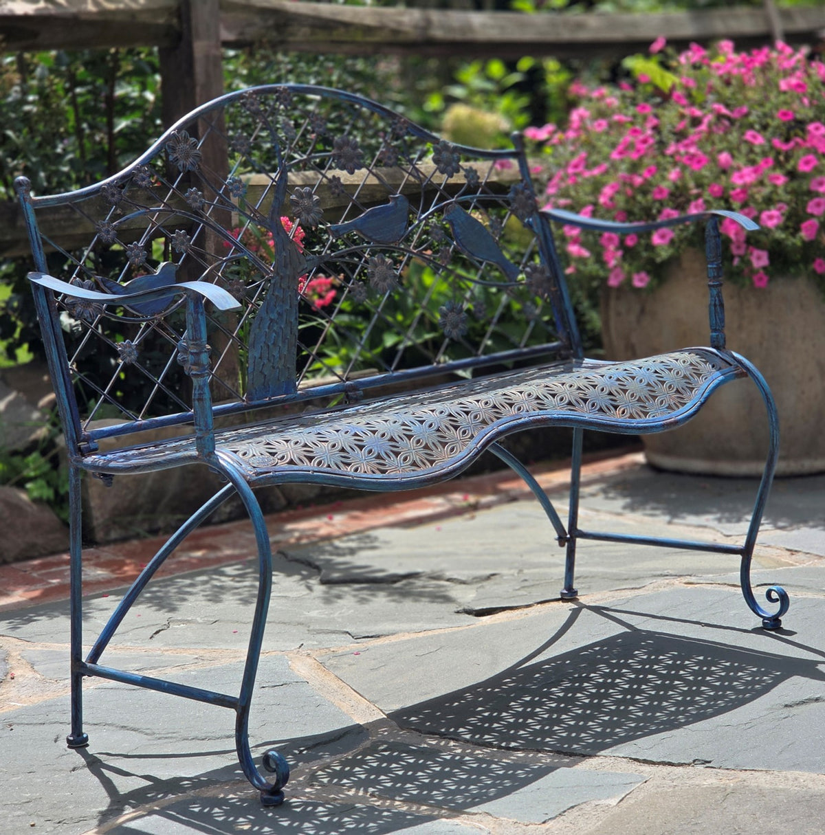 Dark metal garden bench with an ornate tree-and-bird backrest and patterned seat on a stone patio.