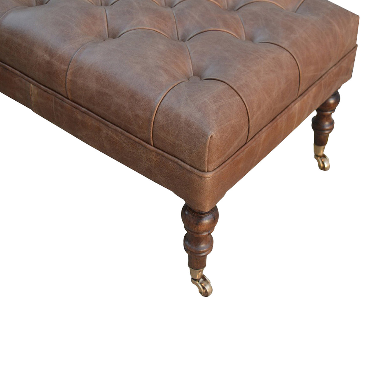 Close-up of the brown leather ottoman's buttoned top corner and turned wooden leg.