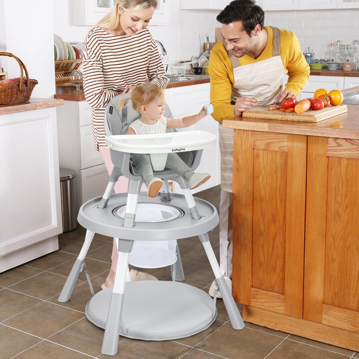 A toddler sits in a gray highchair as two adults prepare food in a kitchen.