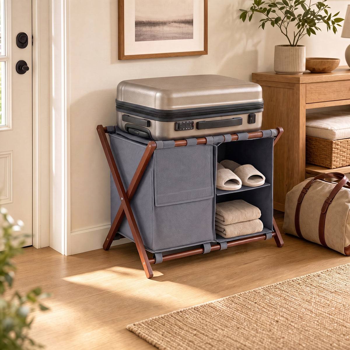 Walnut-toned luggage rack with gray compartments holds a suitcase and folded towels in a bright guest room.