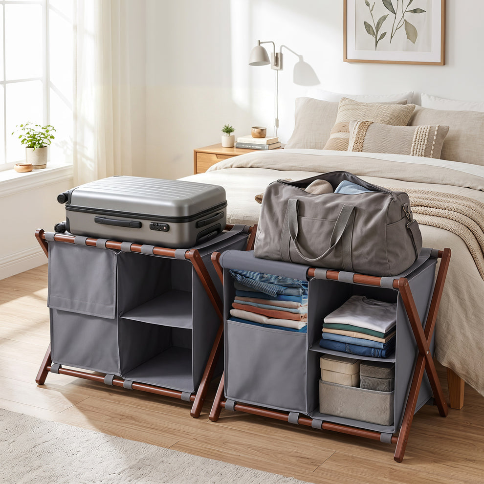 Two gray fabric luggage racks with brown frames hold a suitcase, duffel bag, folded clothes, and storage bins in a bedroom.