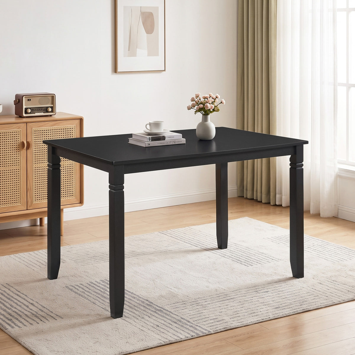 Black rectangular table standing alone on a rug beside a cane-front cabinet and a bright window.