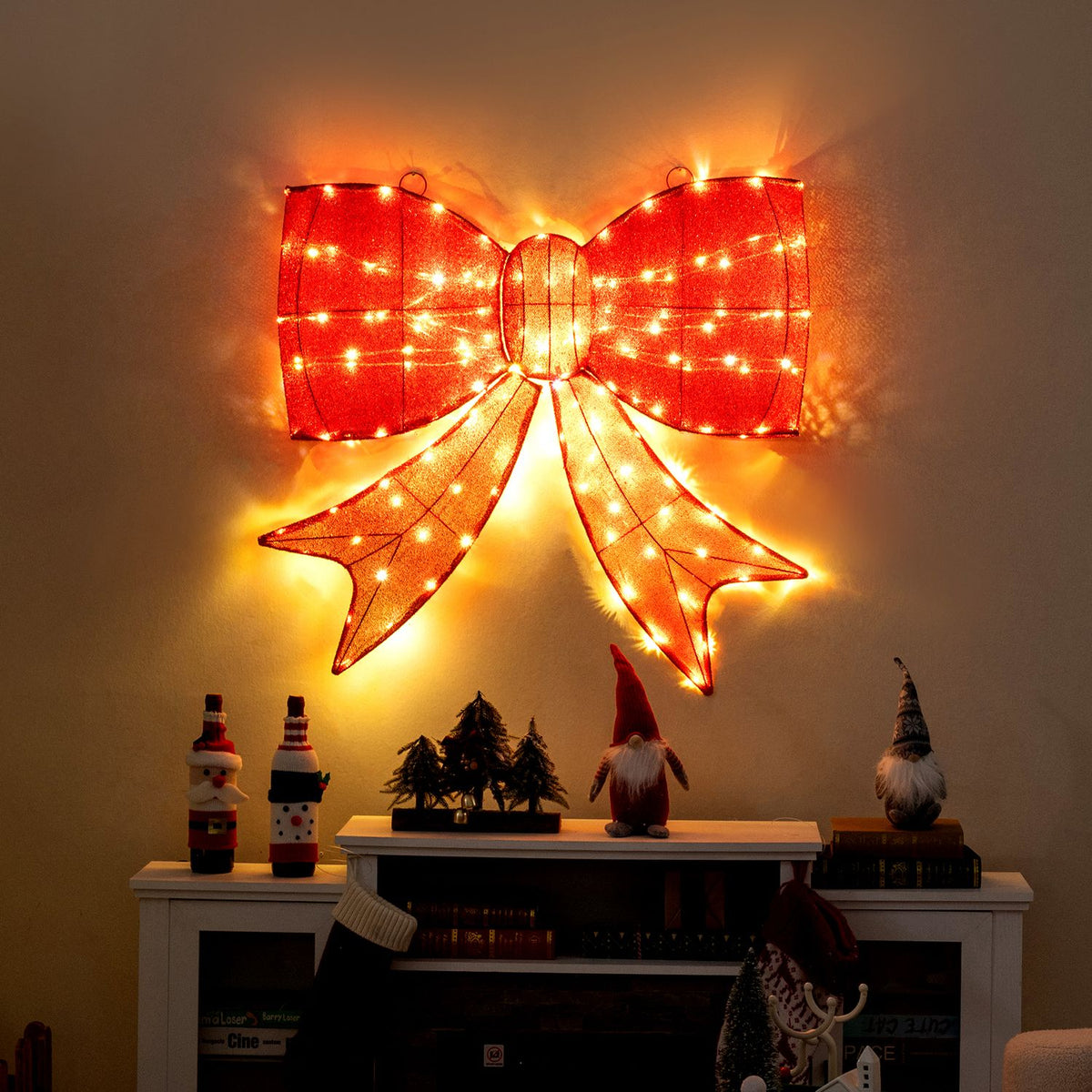 A glowing red bow hangs above a mantel decorated with small holiday figures and trees.