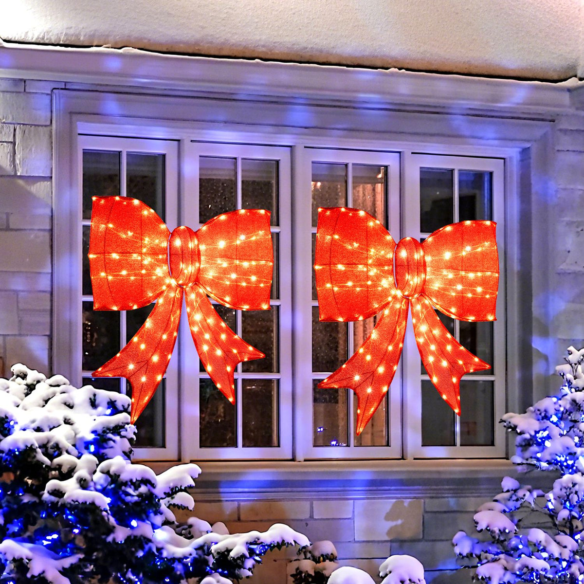 Two glowing red bows hang on a house window above snow-covered shrubs.