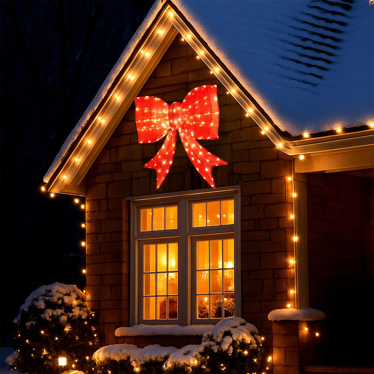 A glowing red bow decorates the gable above a lit window on a snow-covered house.