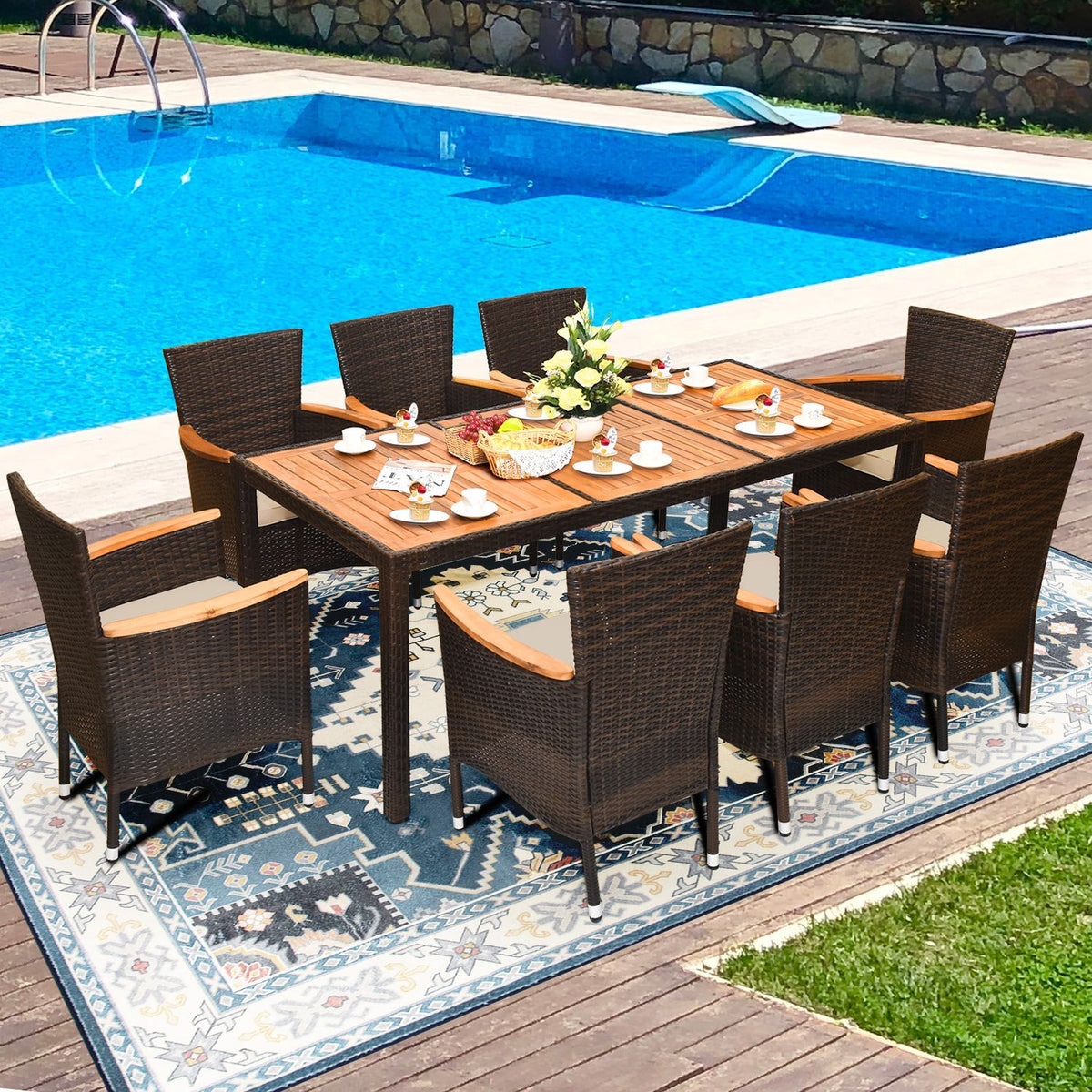 A set dining table with woven chairs stands on a patterned rug beside a swimming pool.