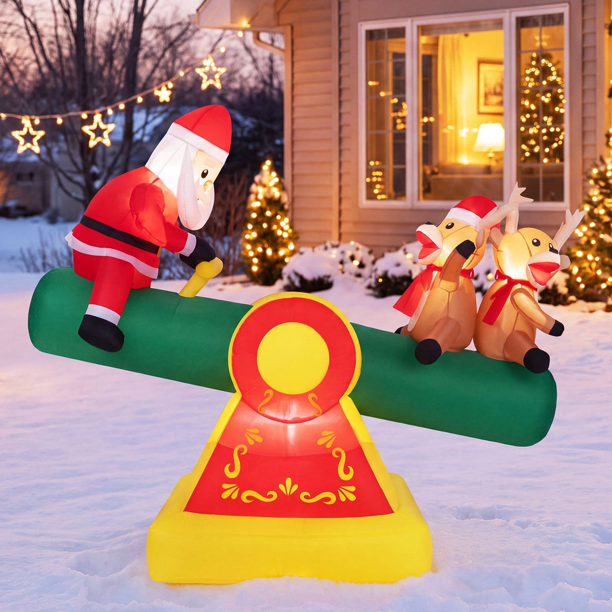 Santa and two reindeer ride a green inflatable teeter-totter in a snowy yard at dusk.