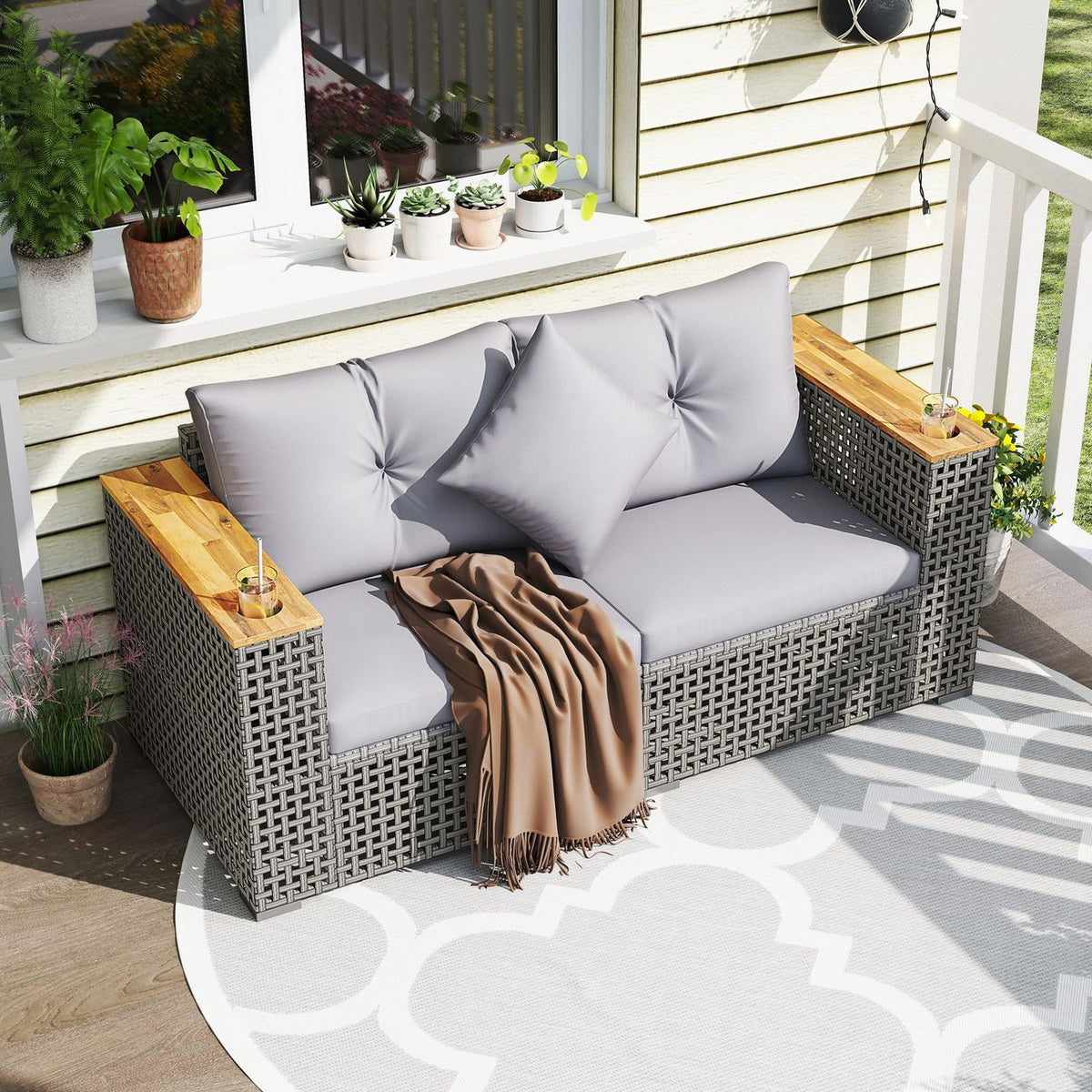 Gray woven loveseat with pale cushions on a patterned patio rug, styled with plants and a draped throw.