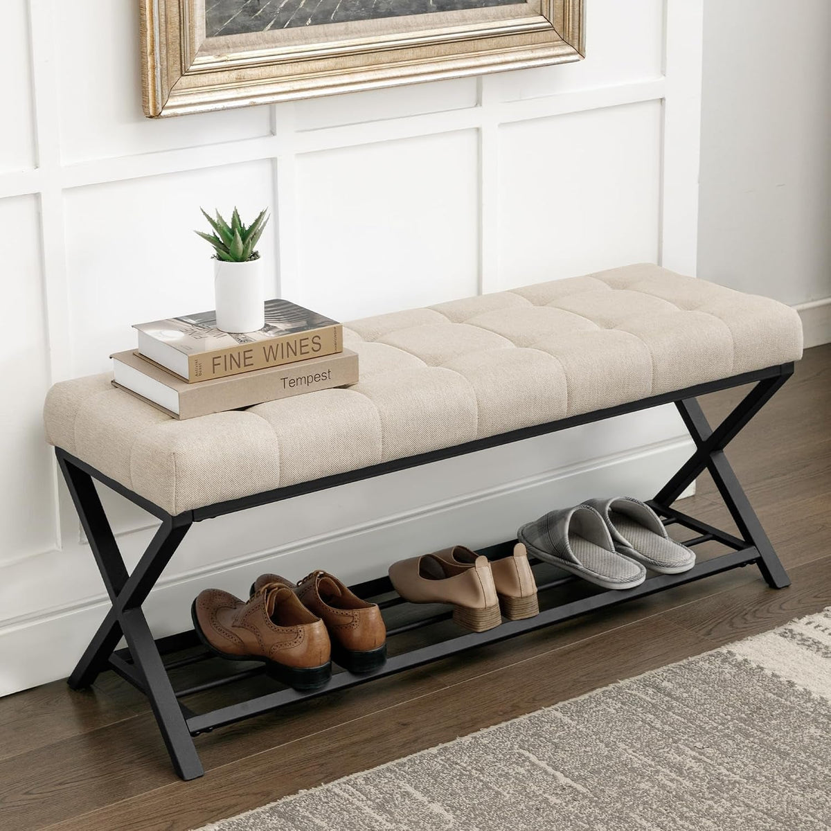 Beige fabric tufted bench showing X-frame legs and books with small plant on top, angled from left