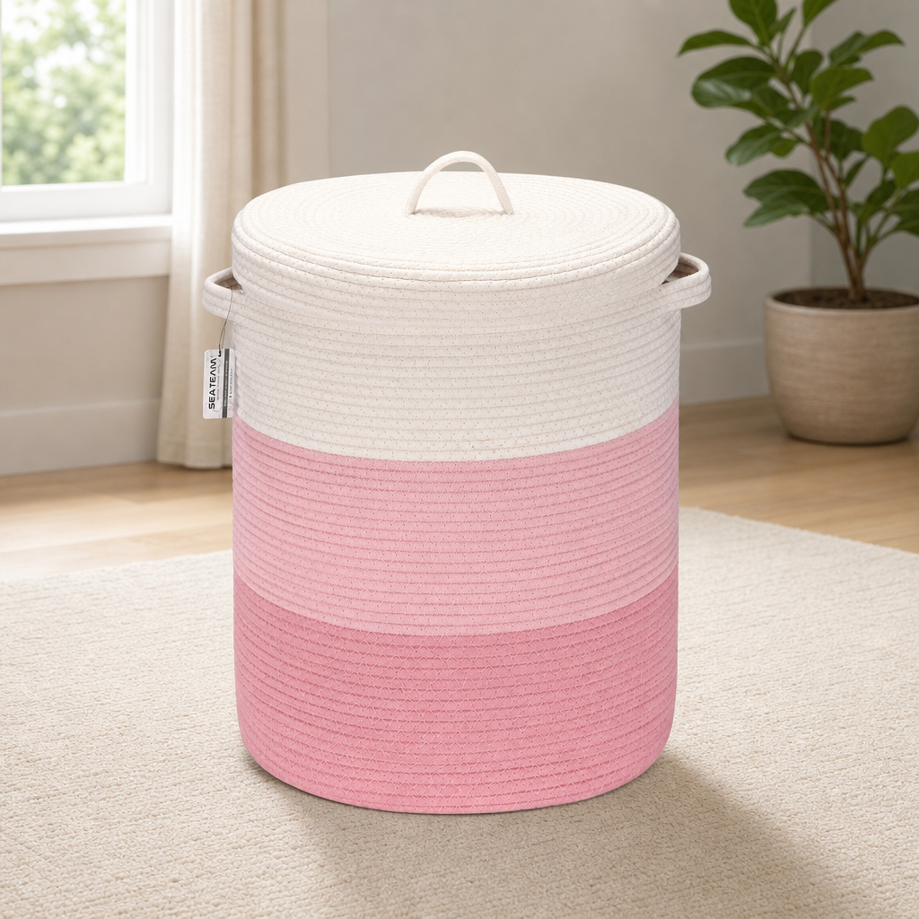 Pink tri‑tone woven cotton rope laundry basket with lid, front view in a bright room next to a plant