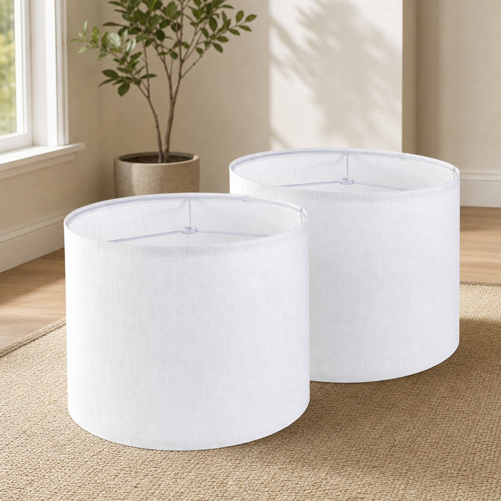 Two white drum lamp shades displayed on a woven rug in a bright room with a potted plant in the background.