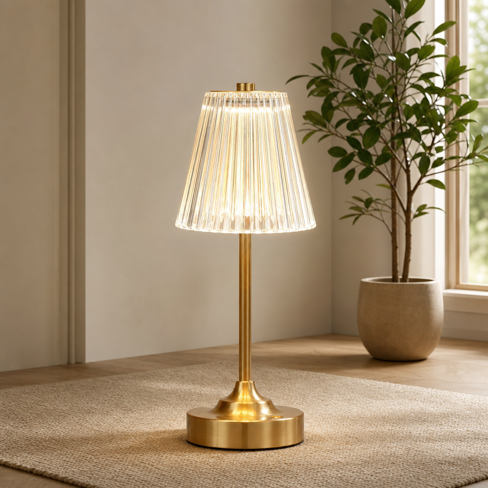 Gold cordless table lamp with clear ribbed acrylic shade, front view on a rug near a potted plant