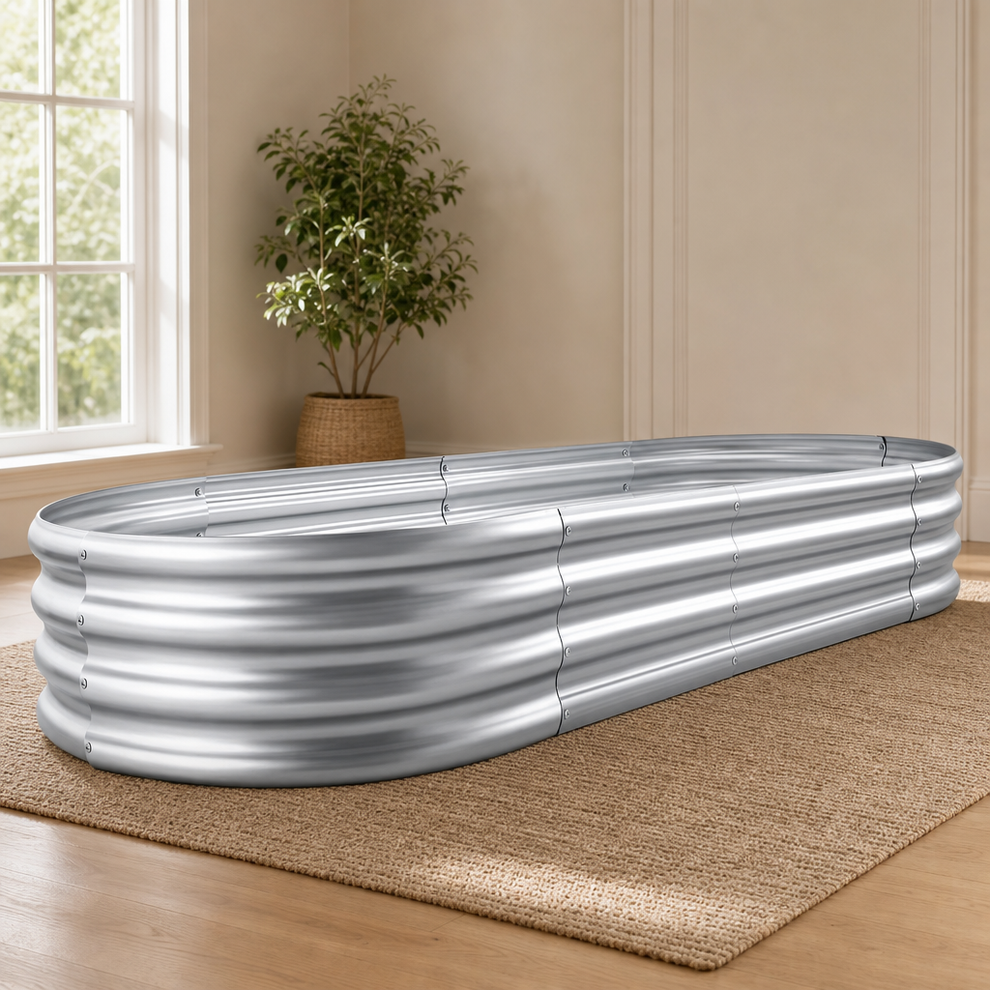 Silver galvanized oval raised garden bed placed on a woven rug near a sunlit window, angled front view
