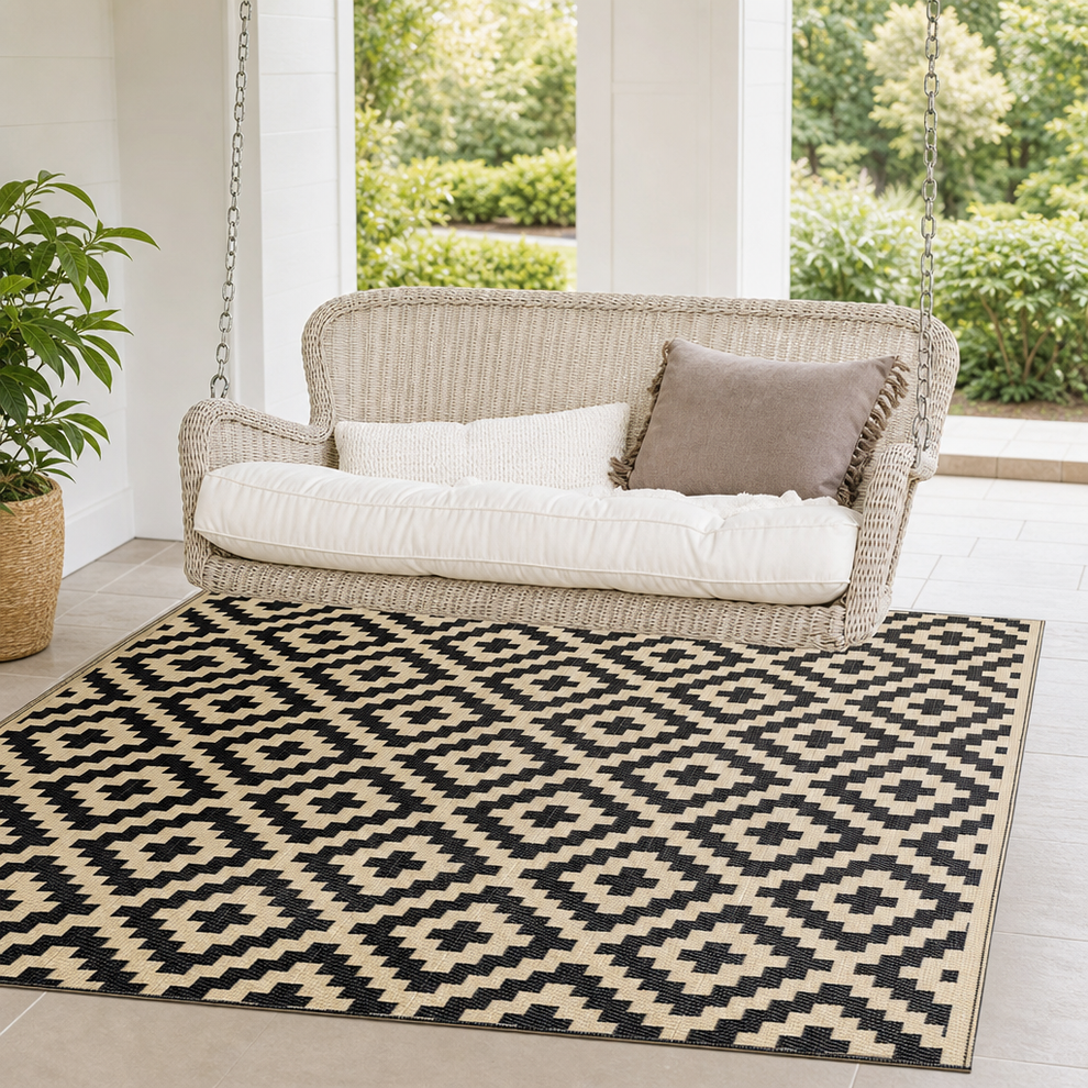 Black and beige geometric flatwoven outdoor rug under a wicker porch swing, angled view