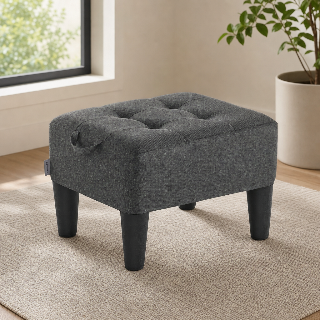 Dark gray linen-look tufted storage ottoman angled front view with tapered black legs in a bright room