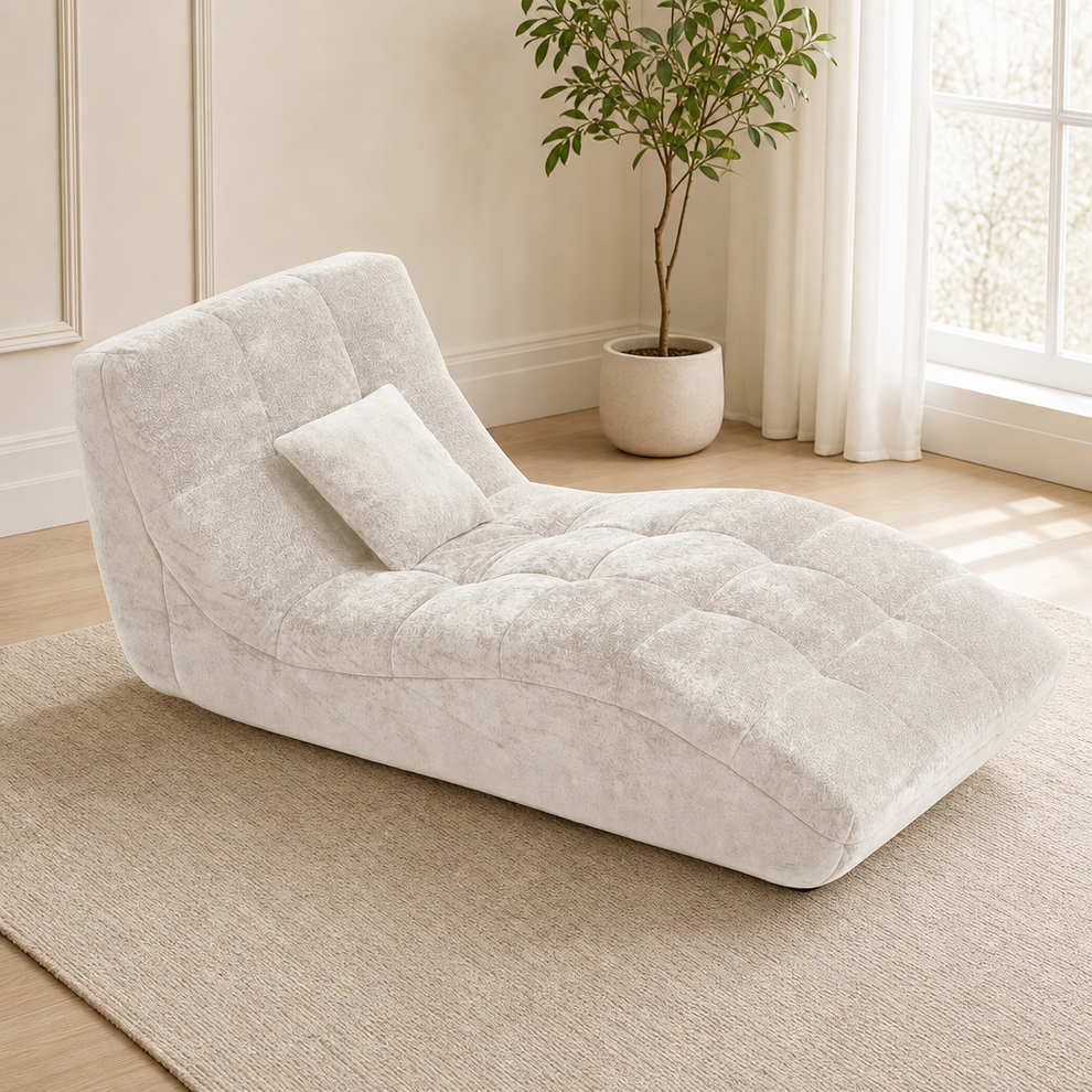 Beige tufted flannel chaise lounge angled front-left view on a rug beside a potted plant and window light