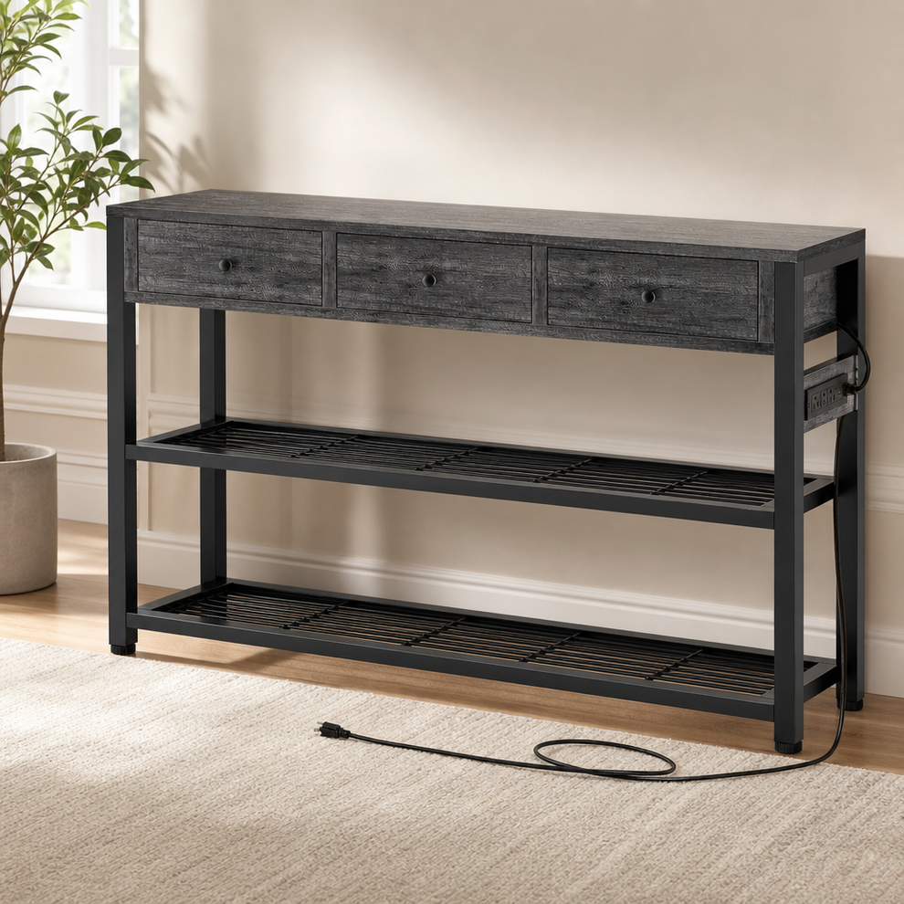 Grey wood-veneer console table with three drawers, black metal frame and two slatted shelves, angled front view with power c