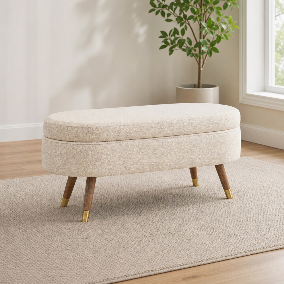 Beige upholstered storage bench angled front view showing wooden legs with gold metal caps in a living room setting