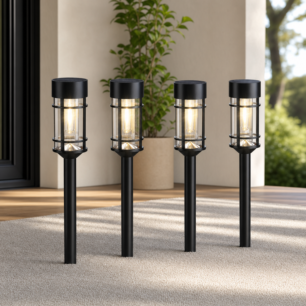Four black solar pathway lights mounted in stakes shown on a patio with plants, front view and warm glow